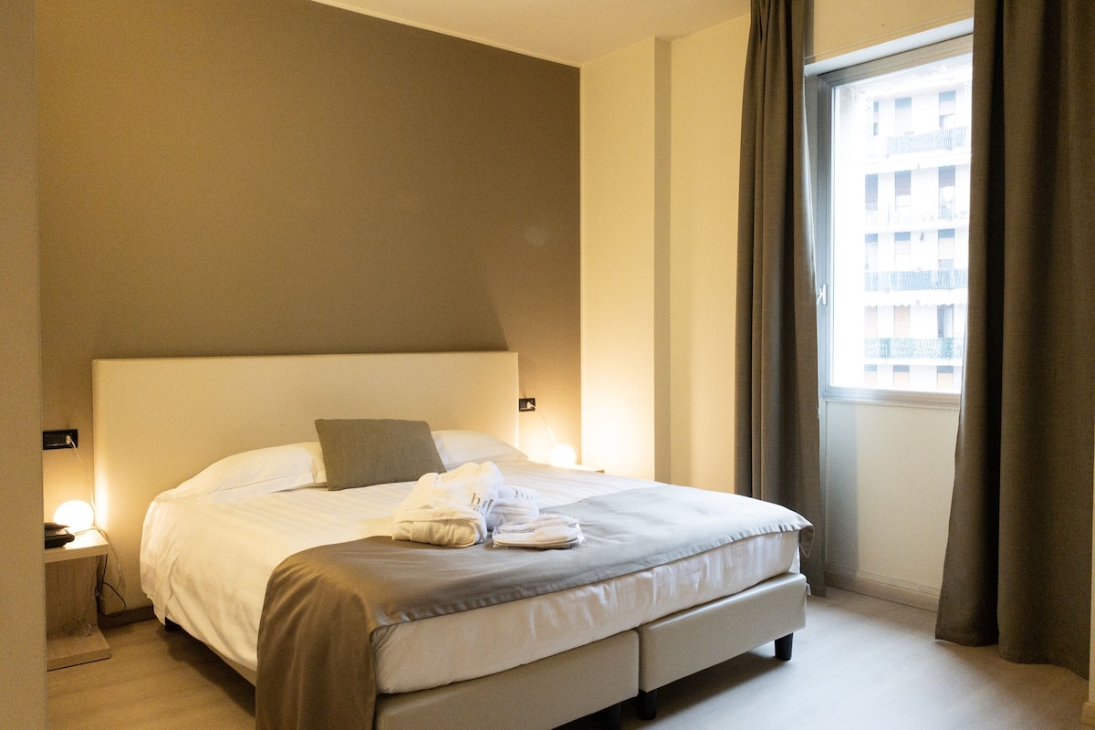 Une chambre d’hôtel avec un grand lit, une table de chevet, une fenêtre avec des rideaux et une vue sur les bâtiments environnants.