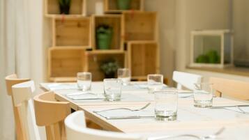 A dining area with a long wooden table set for four, surrounded by white chairs. There are multiple wooden shelves with potted plants in the background.
