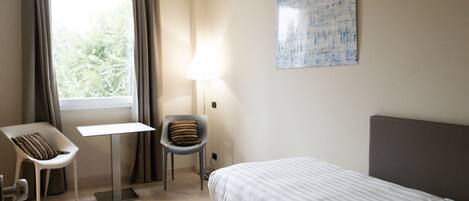 A hotel room with a bed, a desk, a chair, a lamp, and a window with curtains.
