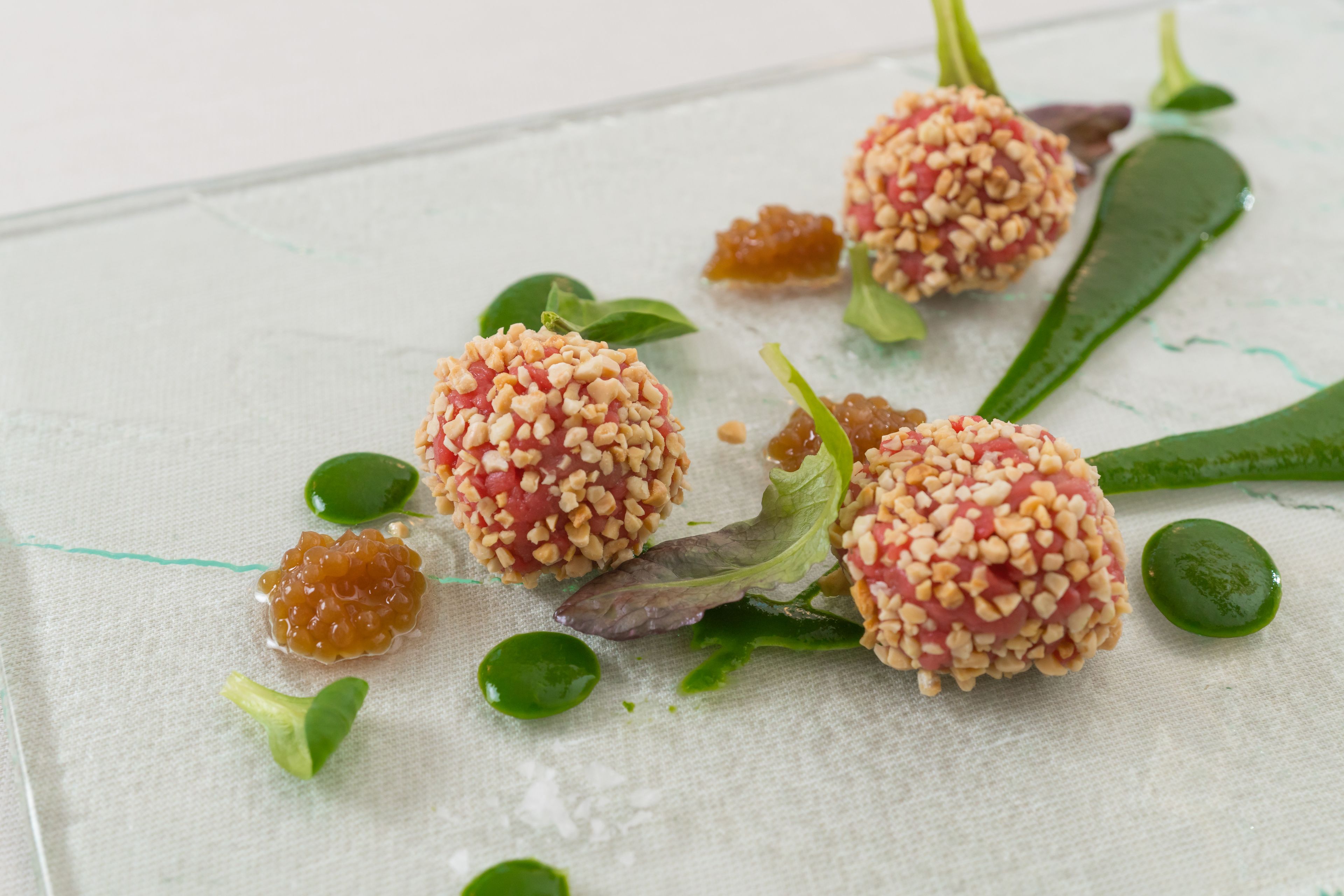 Three spherical food items with a nutty coating, surrounded by green leaves and a white cloth.