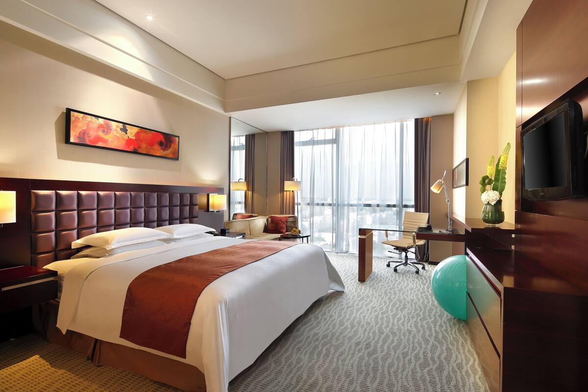 Ein Hotelzimmer mit einem großen Bett, einem Schreibtisch, einem Stuhl, einem Fernseher und einem Fenster mit Vorhängen.