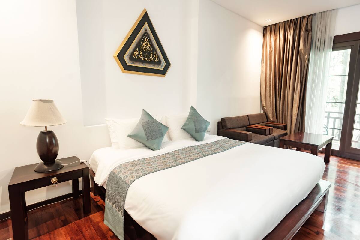Une chambre d’hôtel avec un grand lit, une table de chevet avec une lampe, un petit canapé, un banc en bois et un élément décoratif mural.