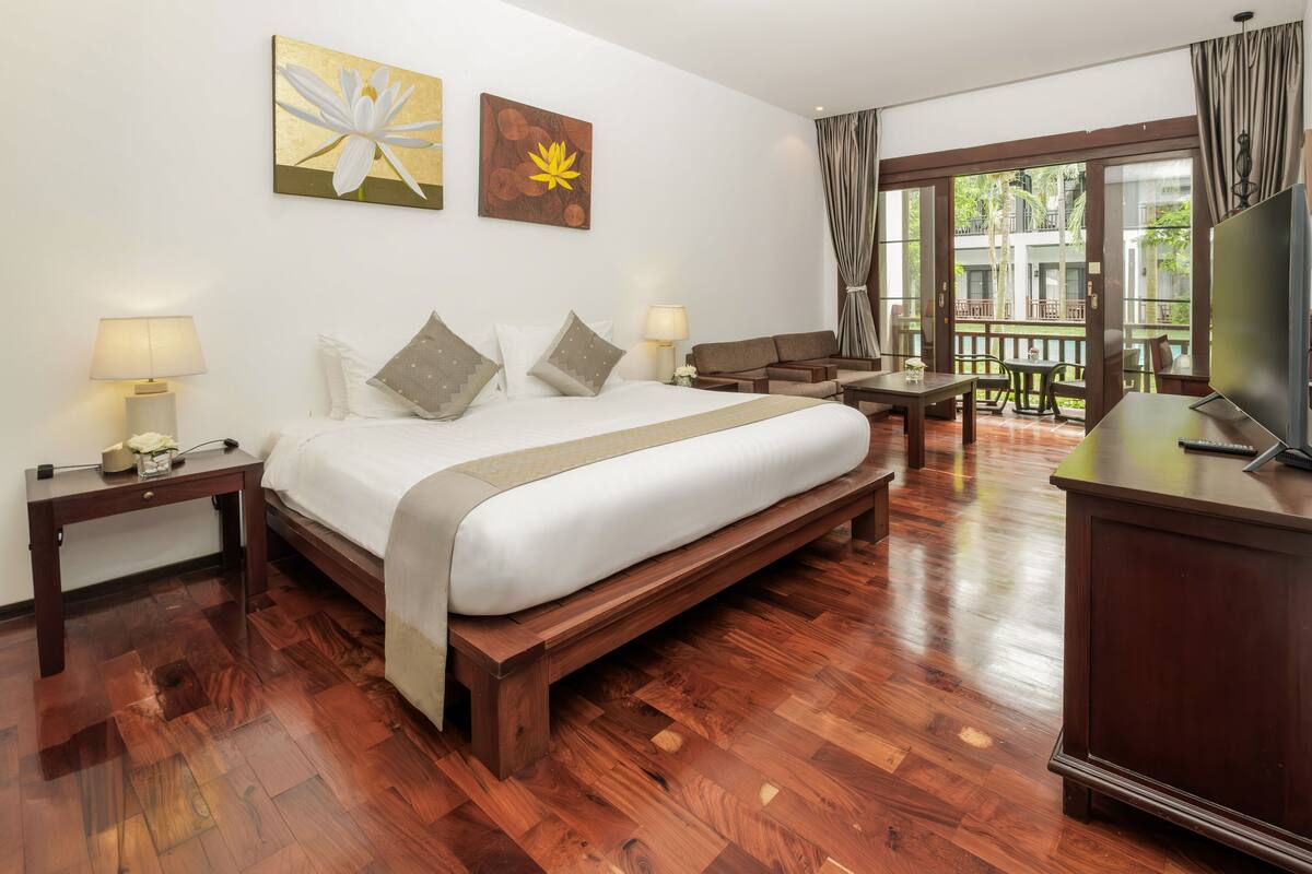 Une chambre d’hôtel avec un grand lit, deux tables de chevet avec des lampes, un bureau en bois, une télévision et un balcon donnant sur l’extérieur.