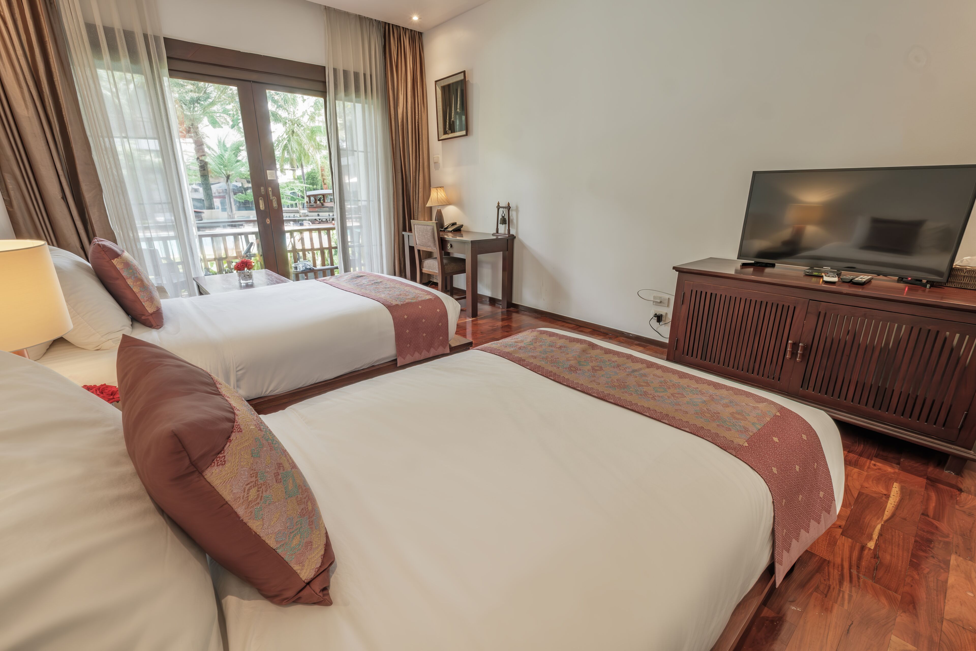 A hotel room with two beds, a television, a desk with a chair, and a view of palm trees through the window.
