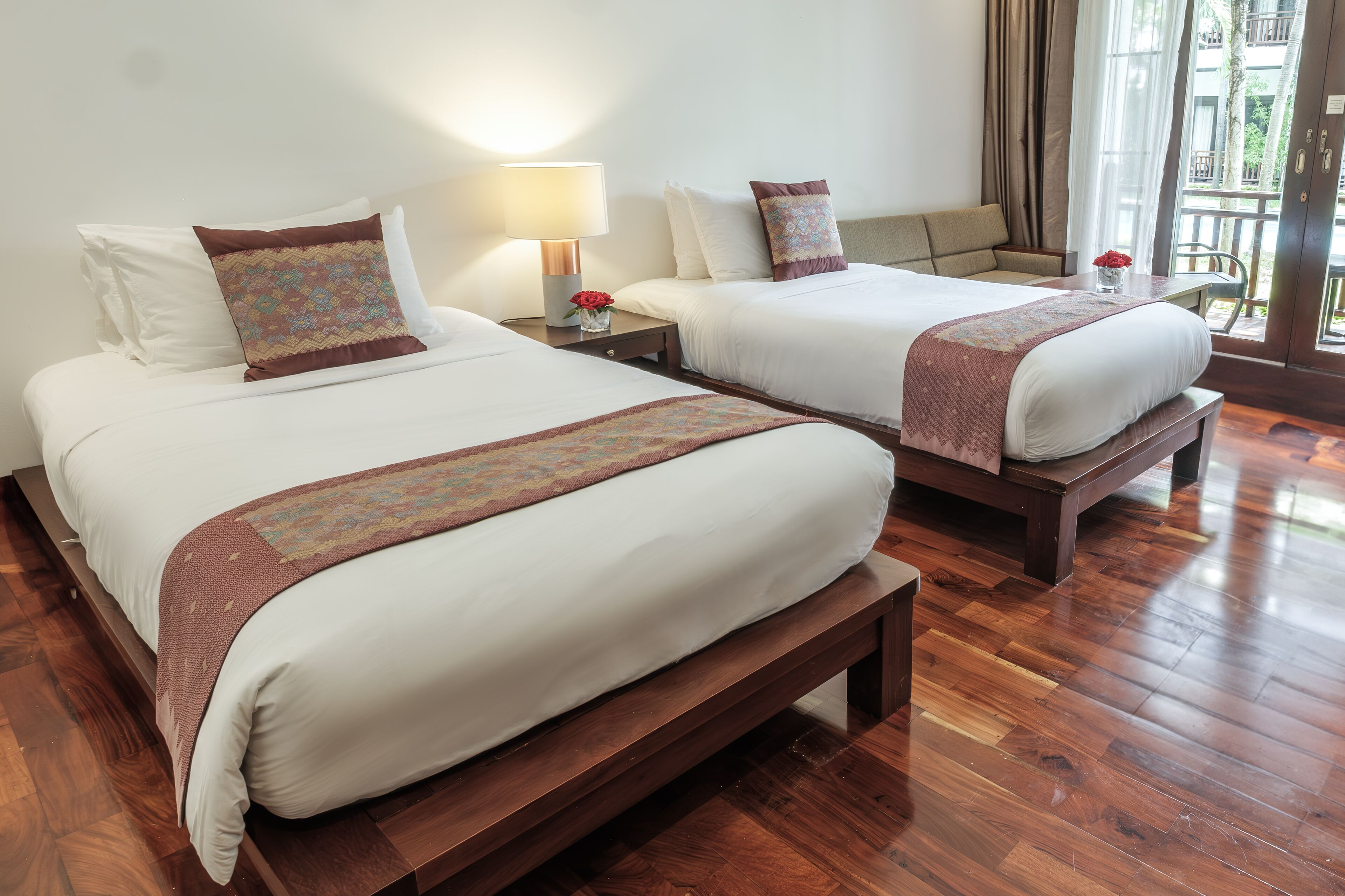 Two double beds with wooden headboards, a bedside table with a lamp, and a window with a view of greenery.