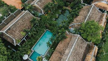 An aerial view of a resort with thatched-roof cottages, a central swimming pool, and lush greenery.