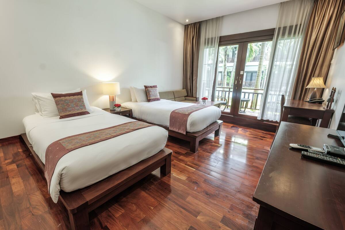 Une chambre d’hôtel avec deux lits, un plancher en bois, un bureau avec une lampe et une vue sur l’extérieur grâce à de grandes fenêtres.