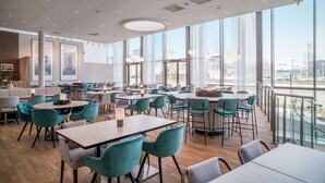 Un espace de restauration moderne avec des tables rondes et des chaises de couleur turquoise, de grandes fenêtres offrant une vue sur la ville et un plancher en bois.