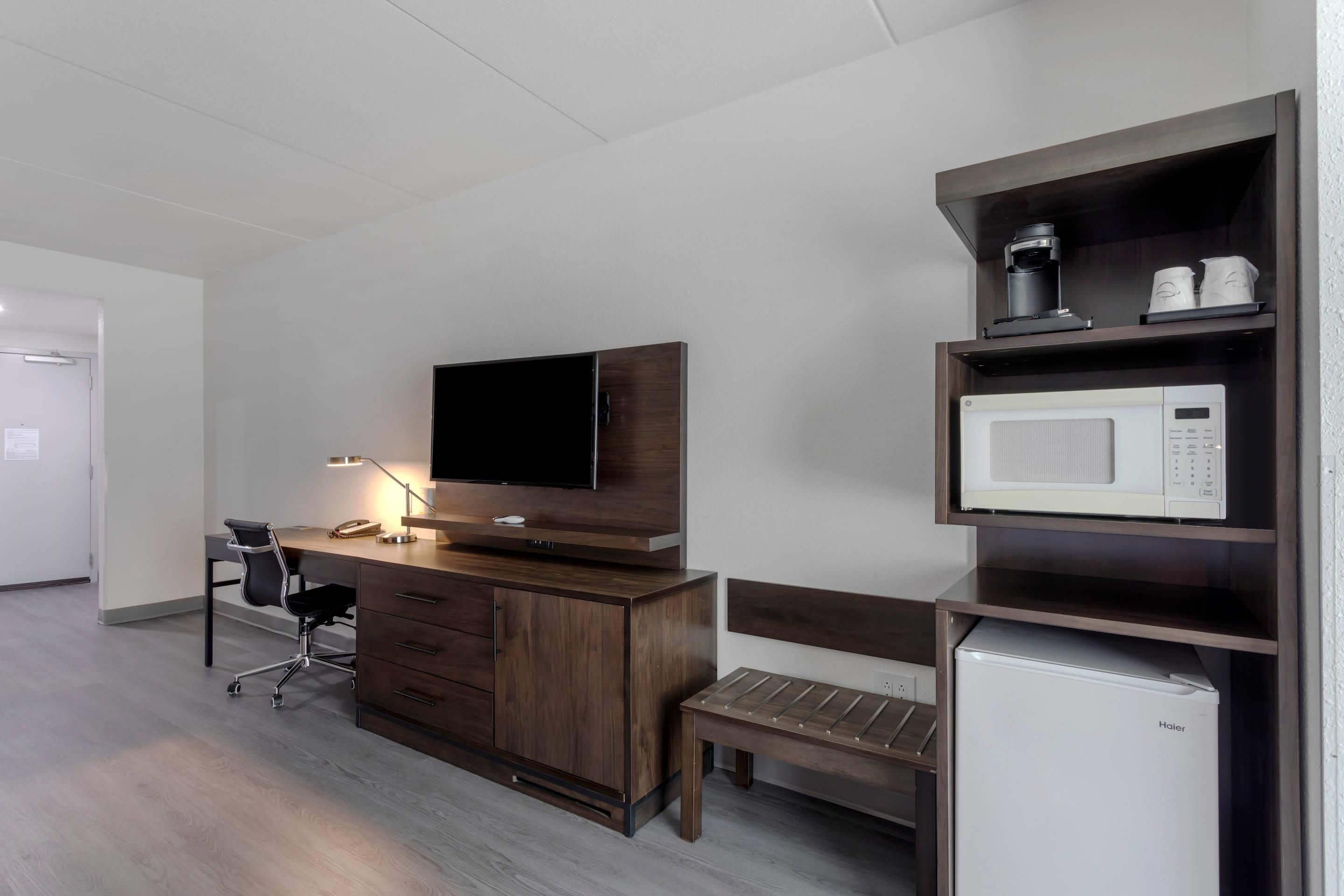 A hotel room with a wooden desk, a television, a microwave, a bench, and a refrigerator.