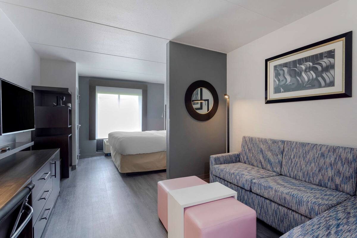 A modern hotel room with a bed, a sofa, a TV, a desk, and a framed artwork on the wall.