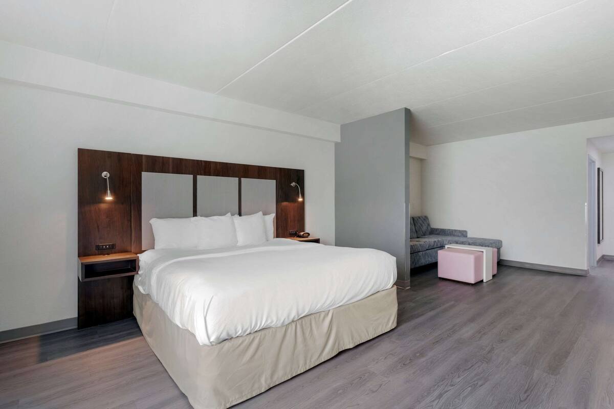 A modern bedroom with a large bed, a nightstand, a bedside table, a grey sofa, and a small pink ottoman.