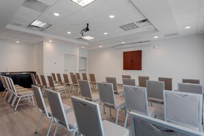 Un salón de conferencias con filas de sillas, un proyector y un panel de madera en la pared.