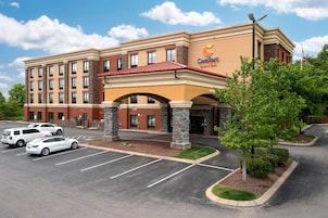 A hotel with a brick facade, a red roof, and a sign that reads 'Comfort Suites.' There are cars parked in front, and a small tree is visible.