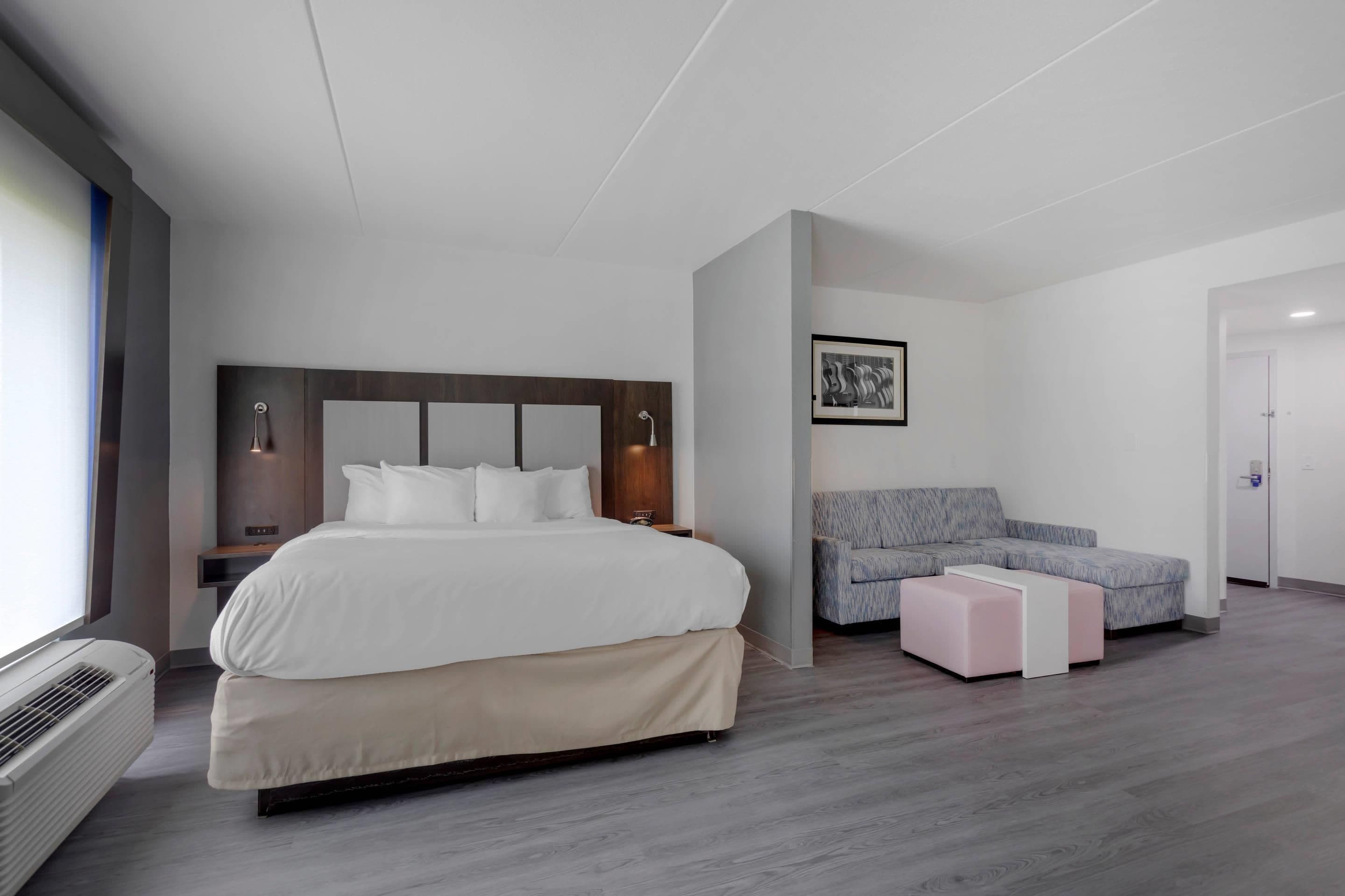 A modern hotel room with a large bed, a grey sofa, a small table, and a framed picture on the wall.