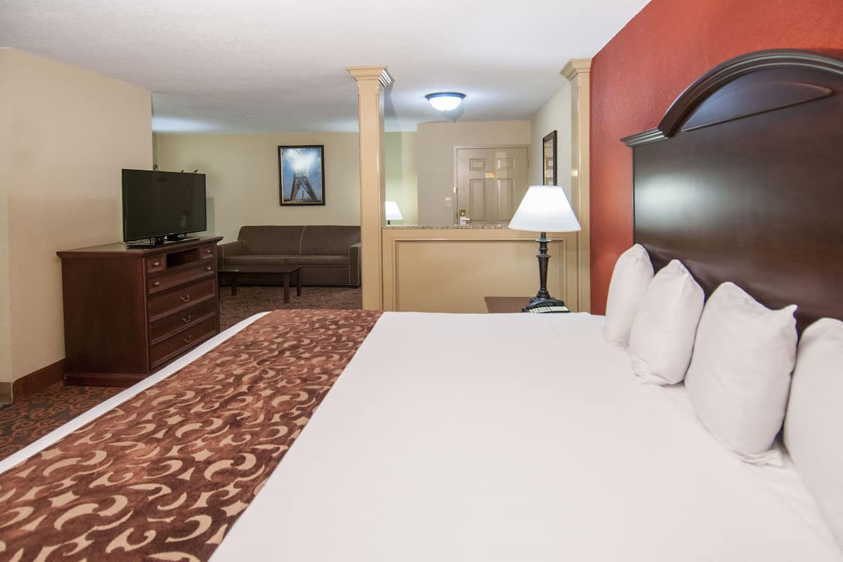 A hotel room with a large bed, a television on a wooden dresser, a sofa, a desk with a lamp, and a mirror.