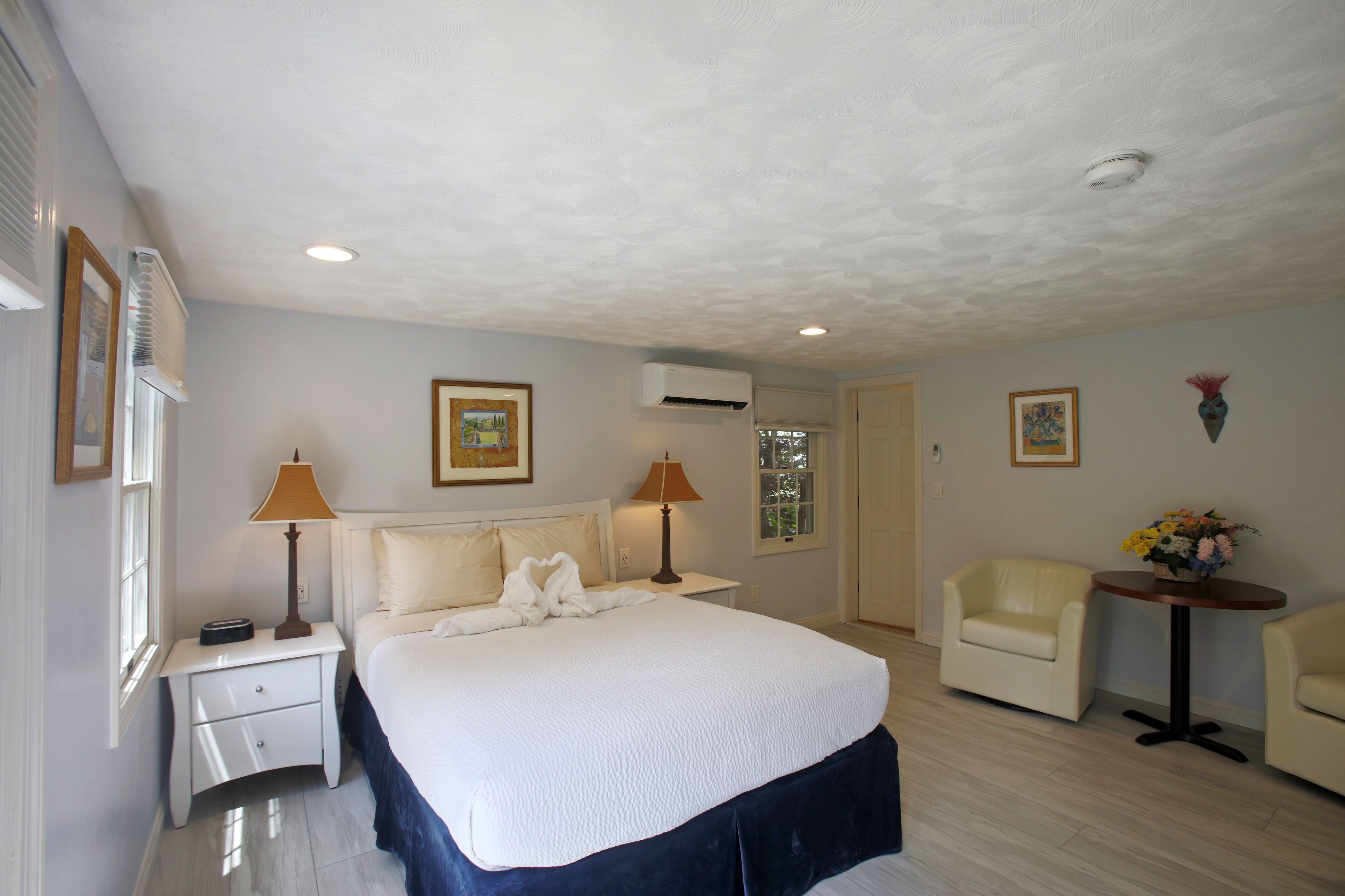 A hotel room with a large bed, two bedside tables with lamps, a chair, a small table with flowers, and an air conditioning unit on the ceiling.
