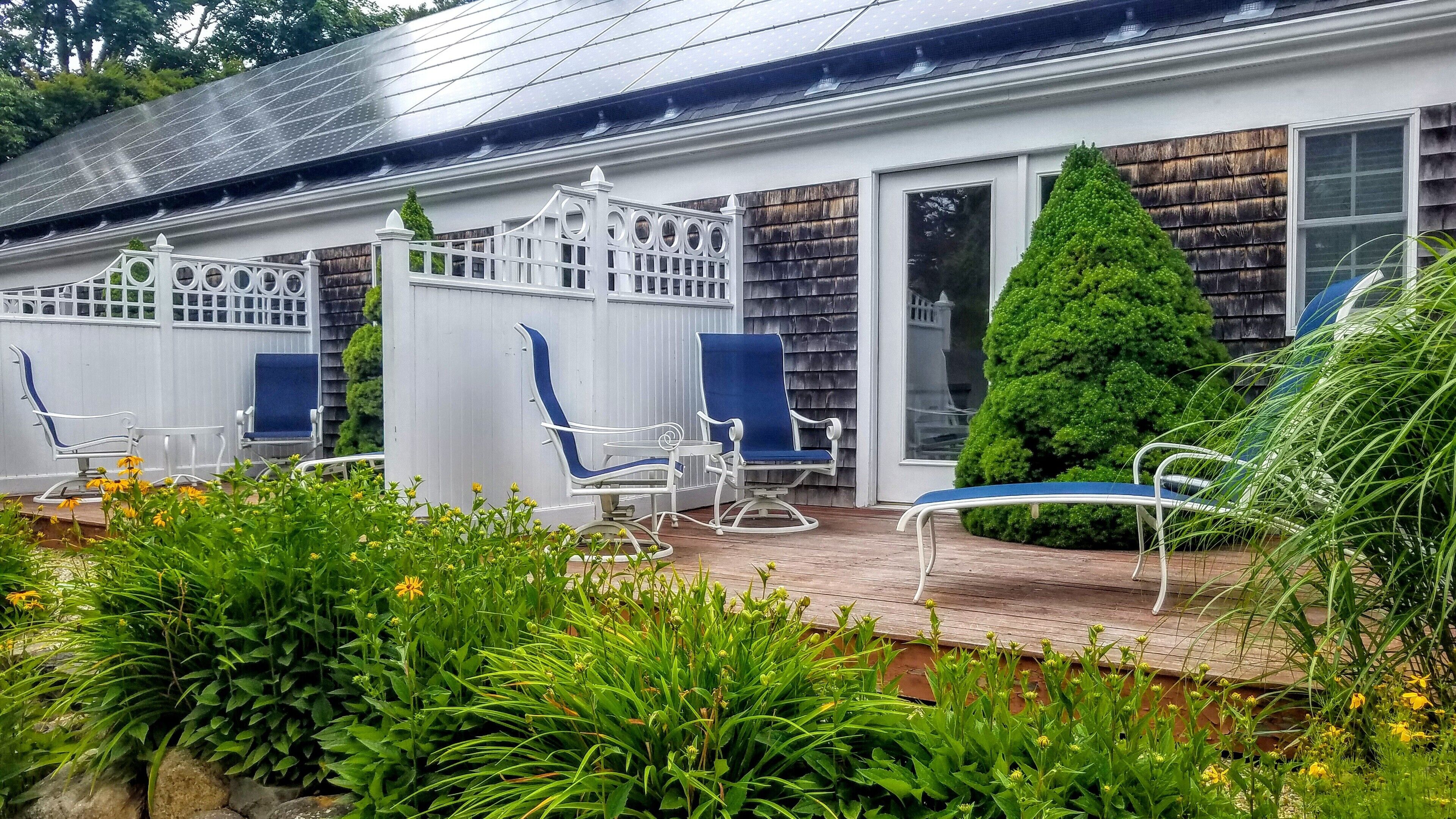 A covered outdoor seating area with white metal chairs and a blue cushioned chair, surrounded by lush greenery and a wooden deck.