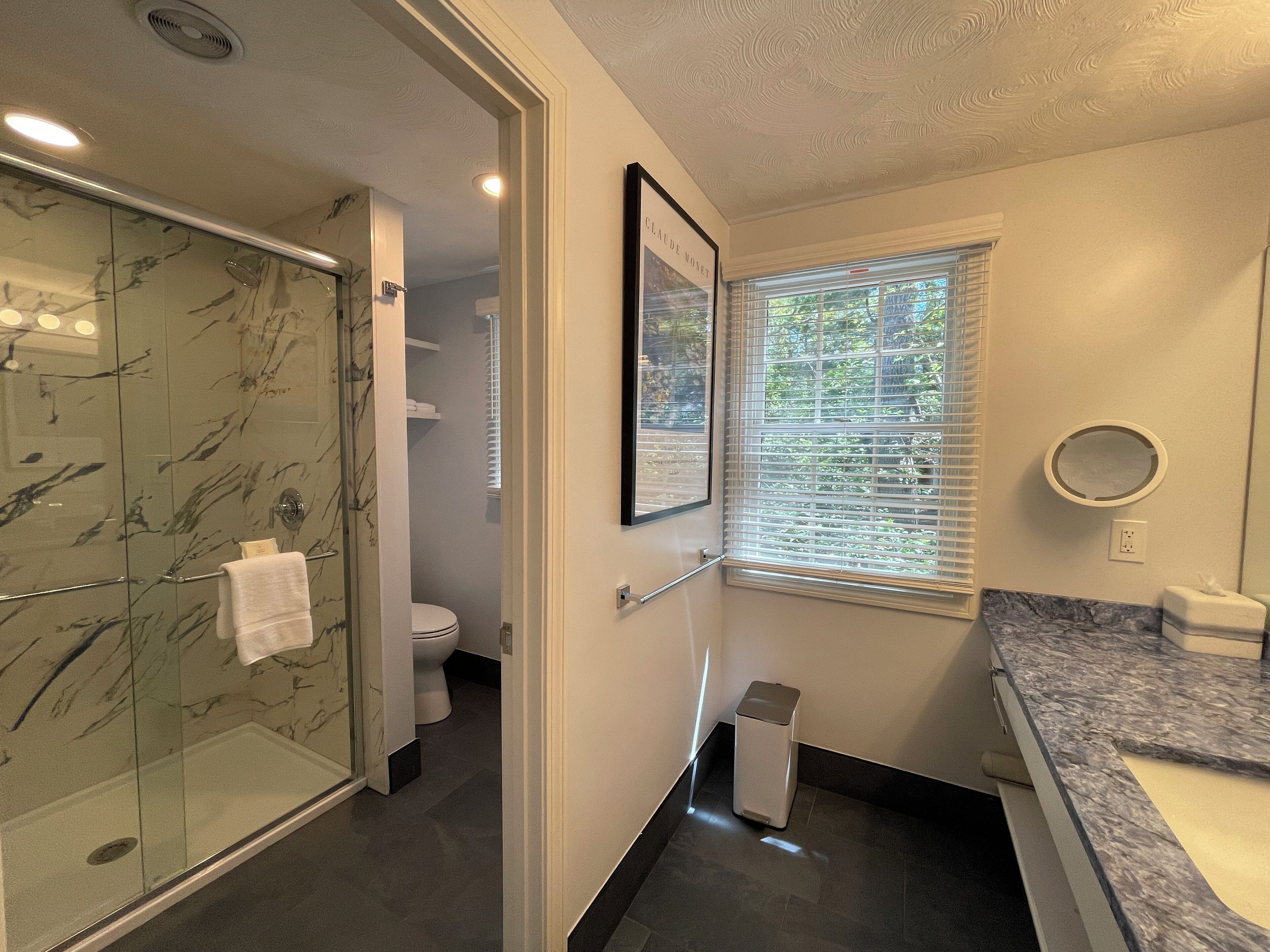 Et moderne badeværelse med en glasbrusekabine, toilet, vask med marmorplade og et vindue med persienner.