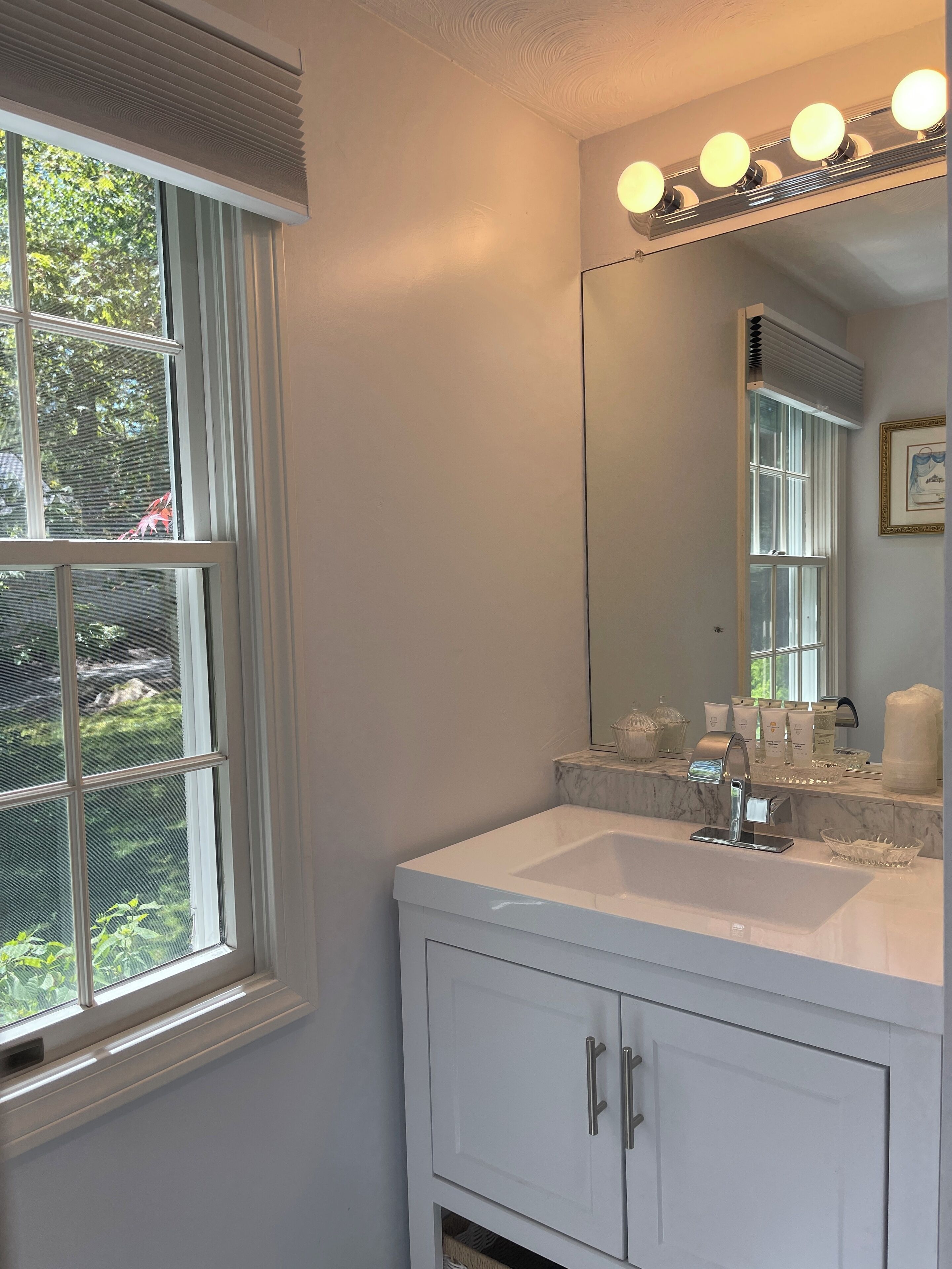 A bathroom with a white vanity, a large mirror with lights, and a window with blinds.