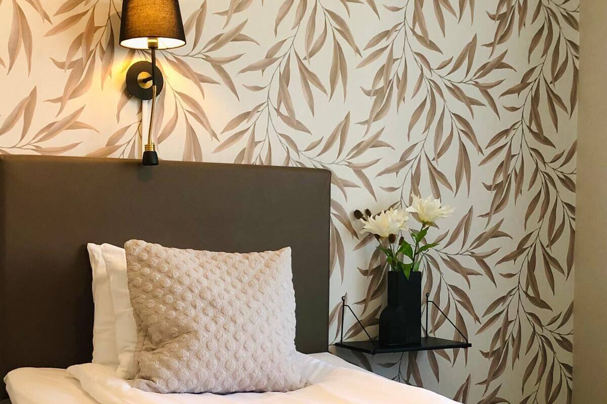 A neatly made bed with a textured white duvet and a beige throw blanket. A wall-mounted lamp with a black shade illuminates the room.