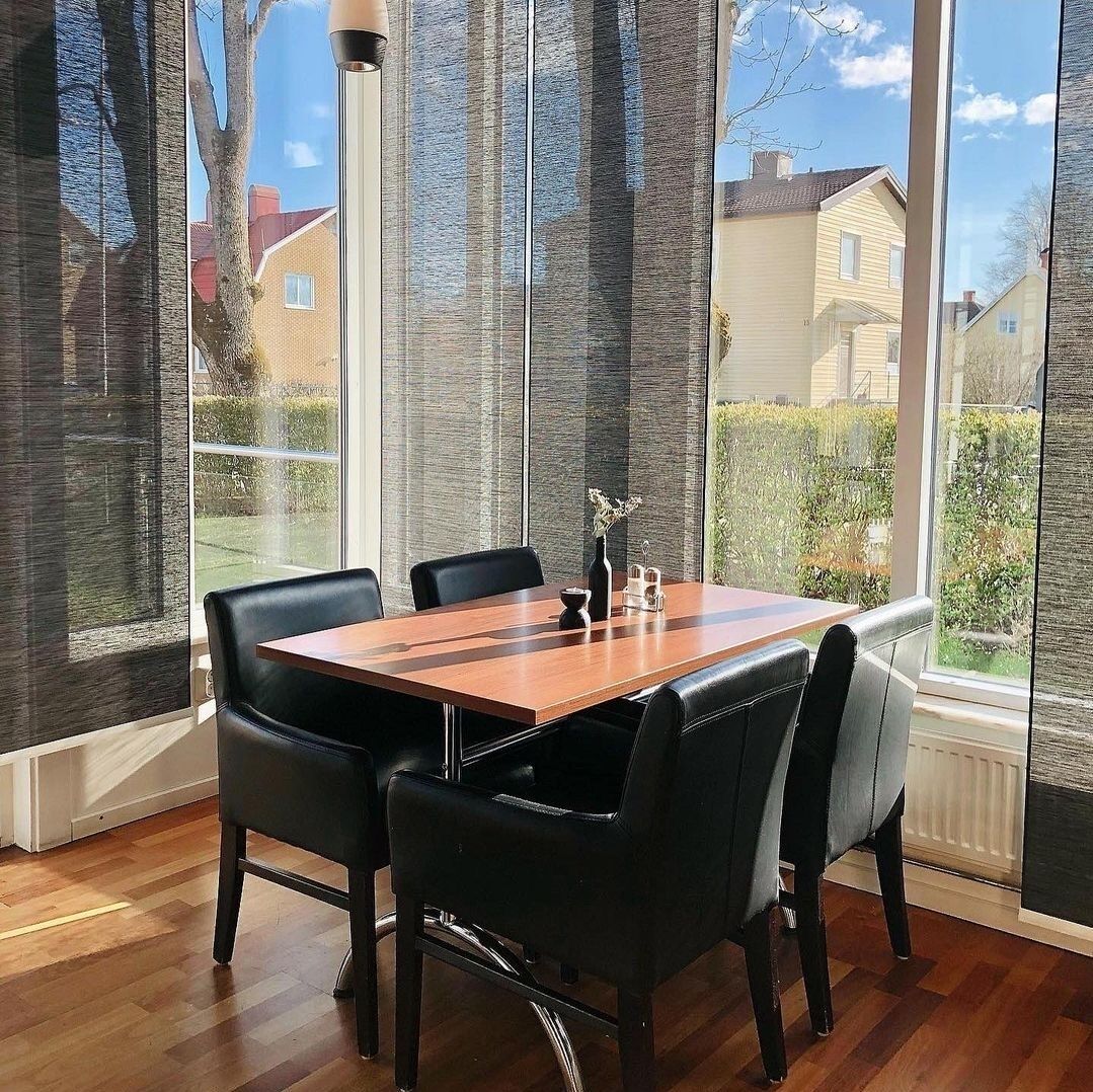 Un comedor con mesa de madera y sillas negras, ventanas amplias con cortinas translúcidas y vista a casas y follaje exterior.