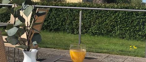 Una mesa de comedor al aire libre con alimentos para el desayuno, un jarrón blanco y una casa de fondo.