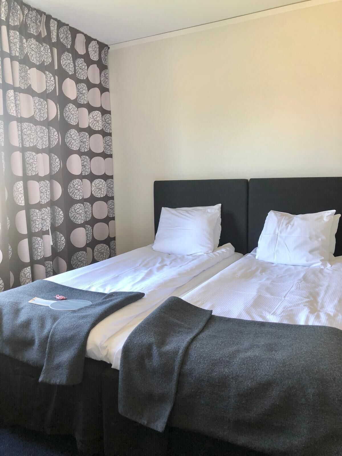 A double bed with white linens, a dark headboard, and a patterned curtain.