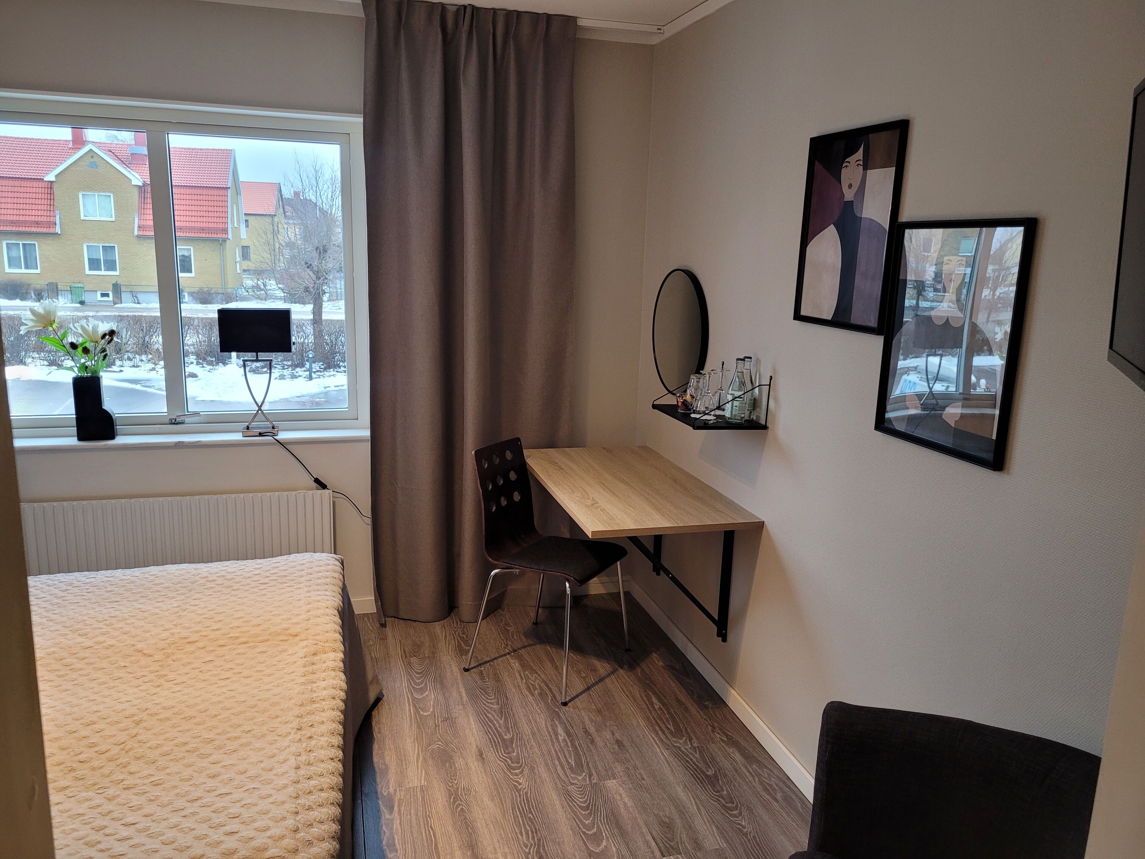 A hotel room with a bed, a desk, a chair, a window with a view of buildings, and a shelf with glasses and a mirror.