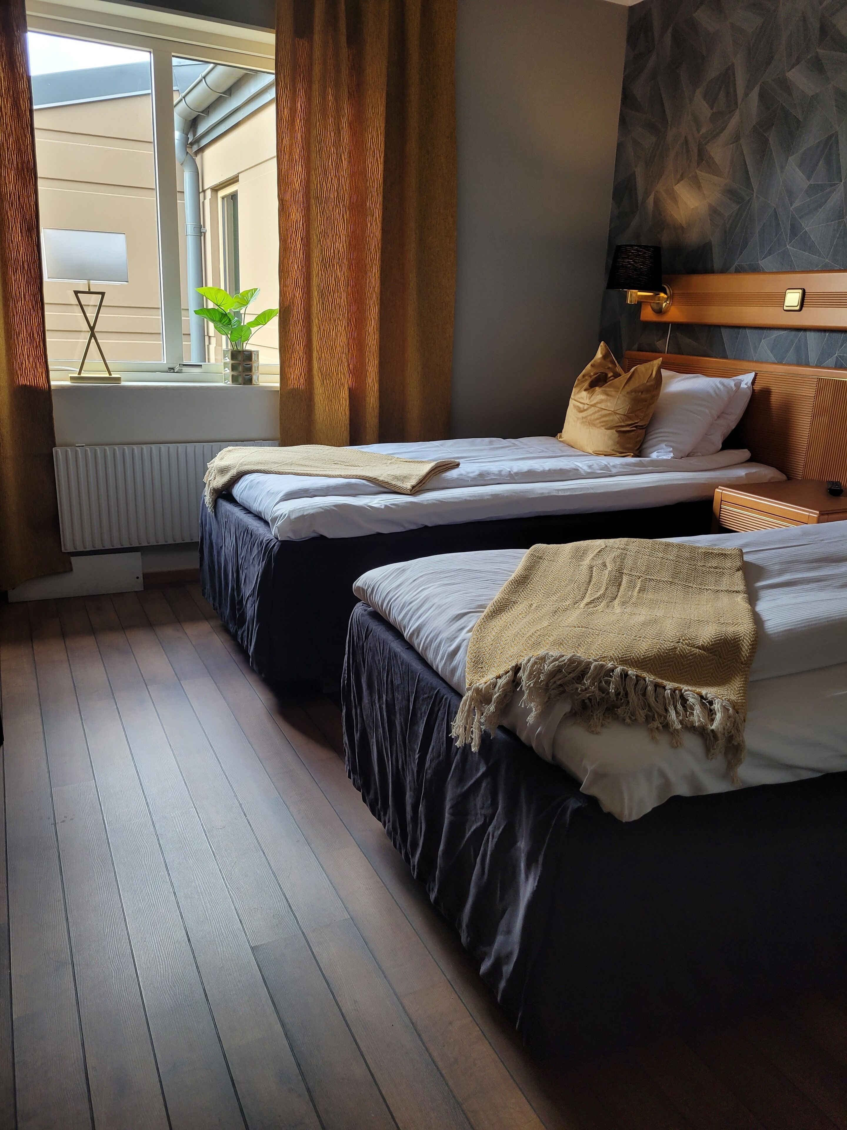 Habitación de hotel con dos camas, una ventana con cortinas, un radiador, una planta y un cabecero de madera.