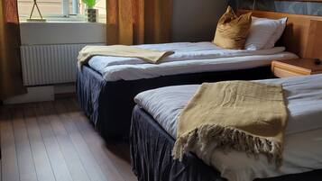 Habitación de hotel con dos camas, una ventana con cortinas, un radiador, una planta y un cabecero de madera.