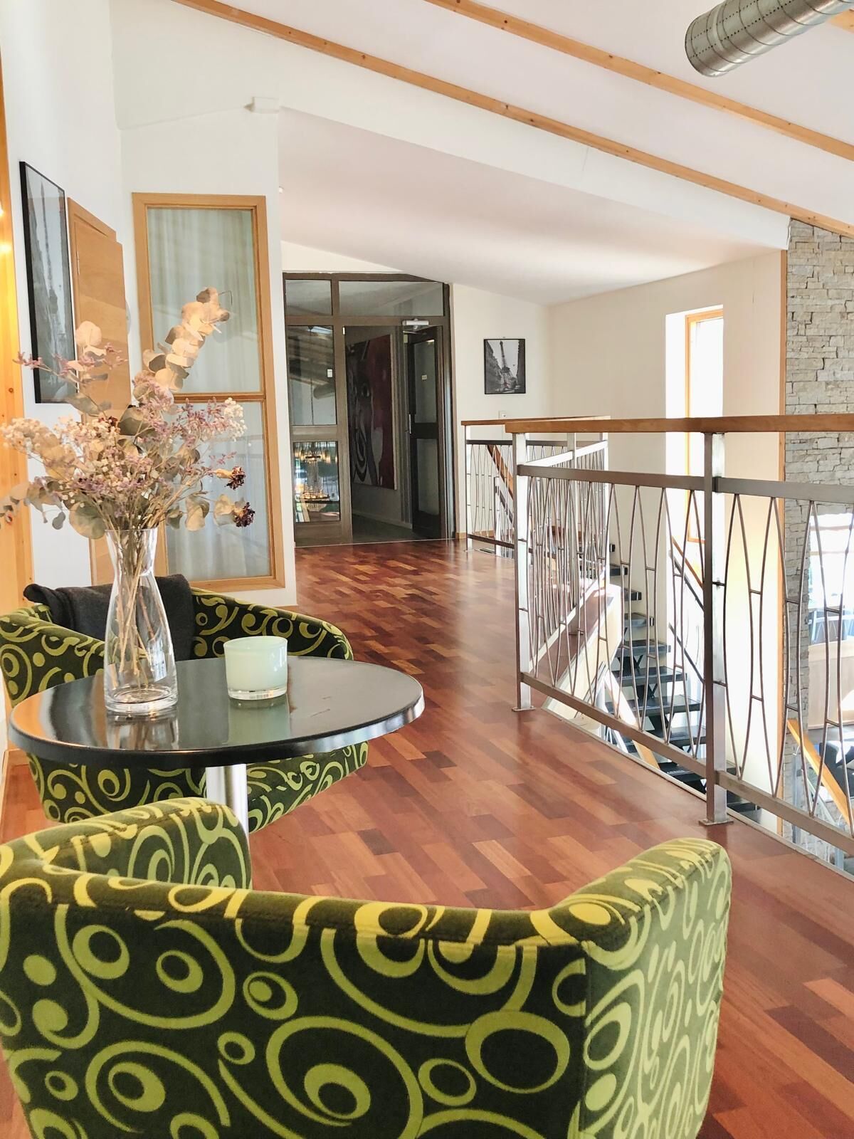 A modern interior with a wooden floor, a glass table, green patterned chairs, and a metal railing overlooking a staircase.