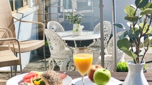 Un área de comedor al aire libre con sillas de mimbre, una mesa con un jarrón blanco y un vaso de jugo de naranja, platos con comida y una taza blanca.