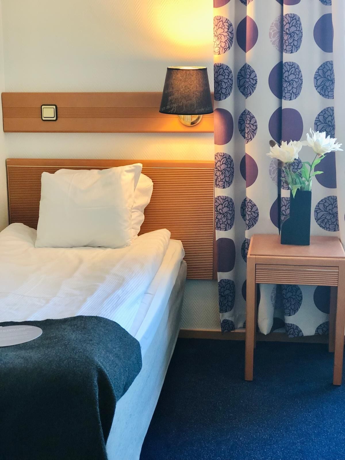 A hotel room with a bed, a bedside lamp, a small table with a vase of flowers, and patterned curtains.