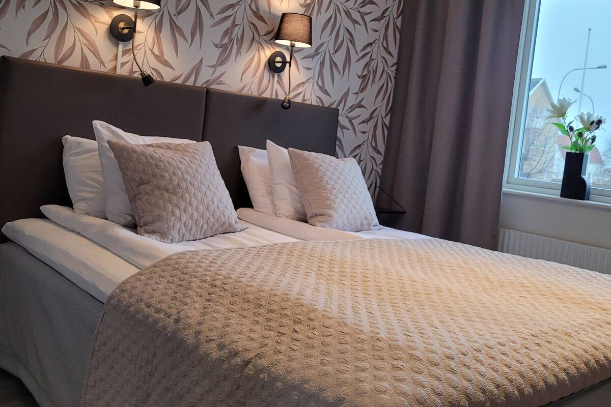 A bedroom with a large bed, two wall-mounted lamps, a window with curtains, and a patterned wallpaper.