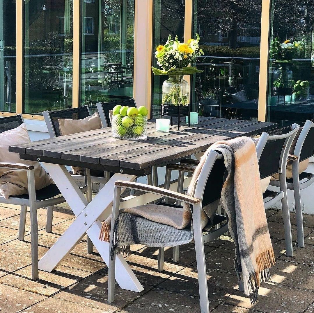 Et udendørs spiseområde med et bord, stole, en blomstervase og en kurv med citroner.