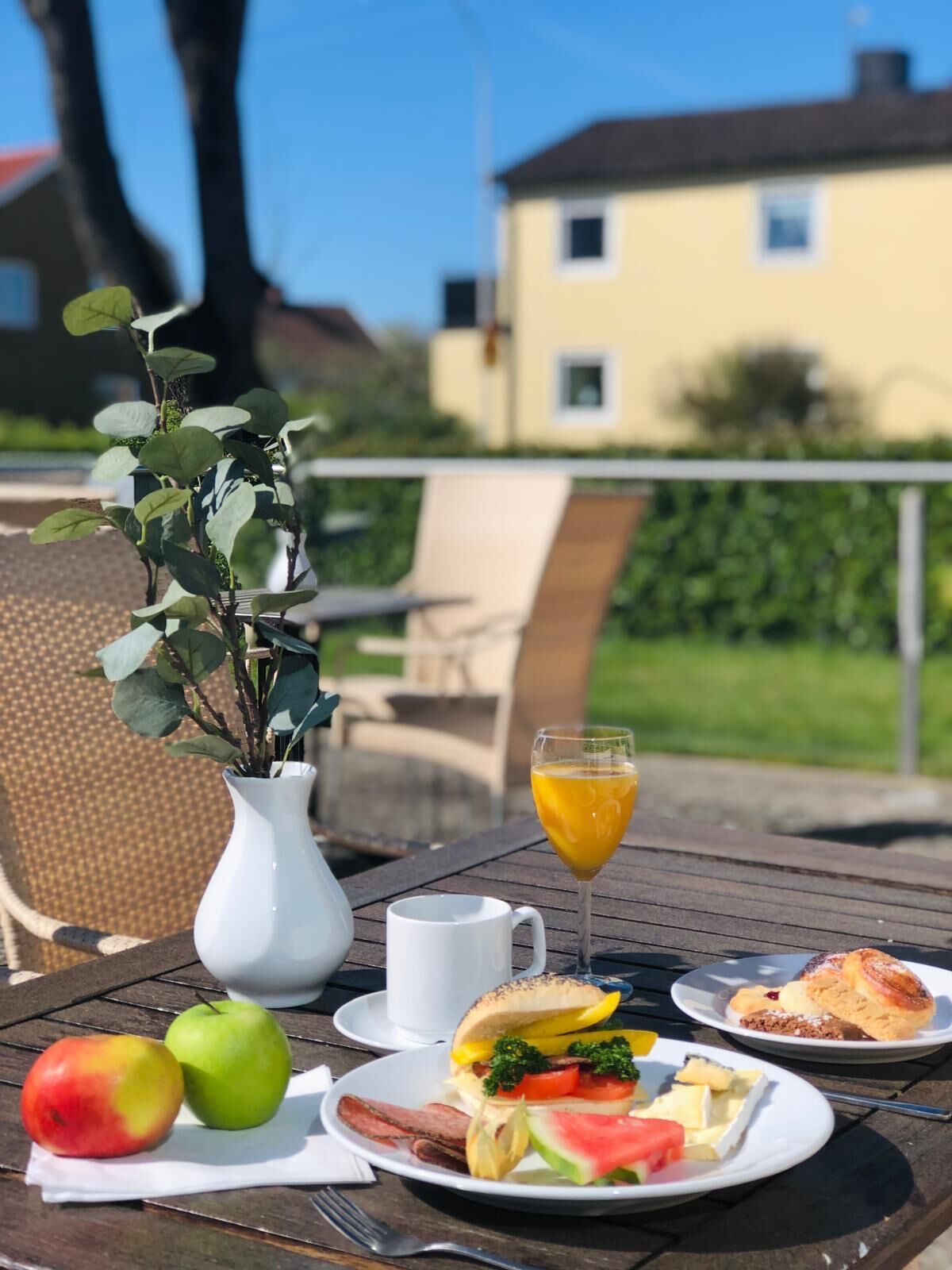 Une table dressée à l’extérieur, avec un repas, un verre de jus d’orange et un vase blanc contenant des feuilles vertes.