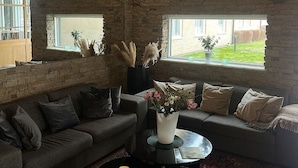 Una sala de estar acogedora con una pared de piedra, un sofá gris, una mesa redonda de centro con velas y vistas a un jardín a través de grandes ventanales.