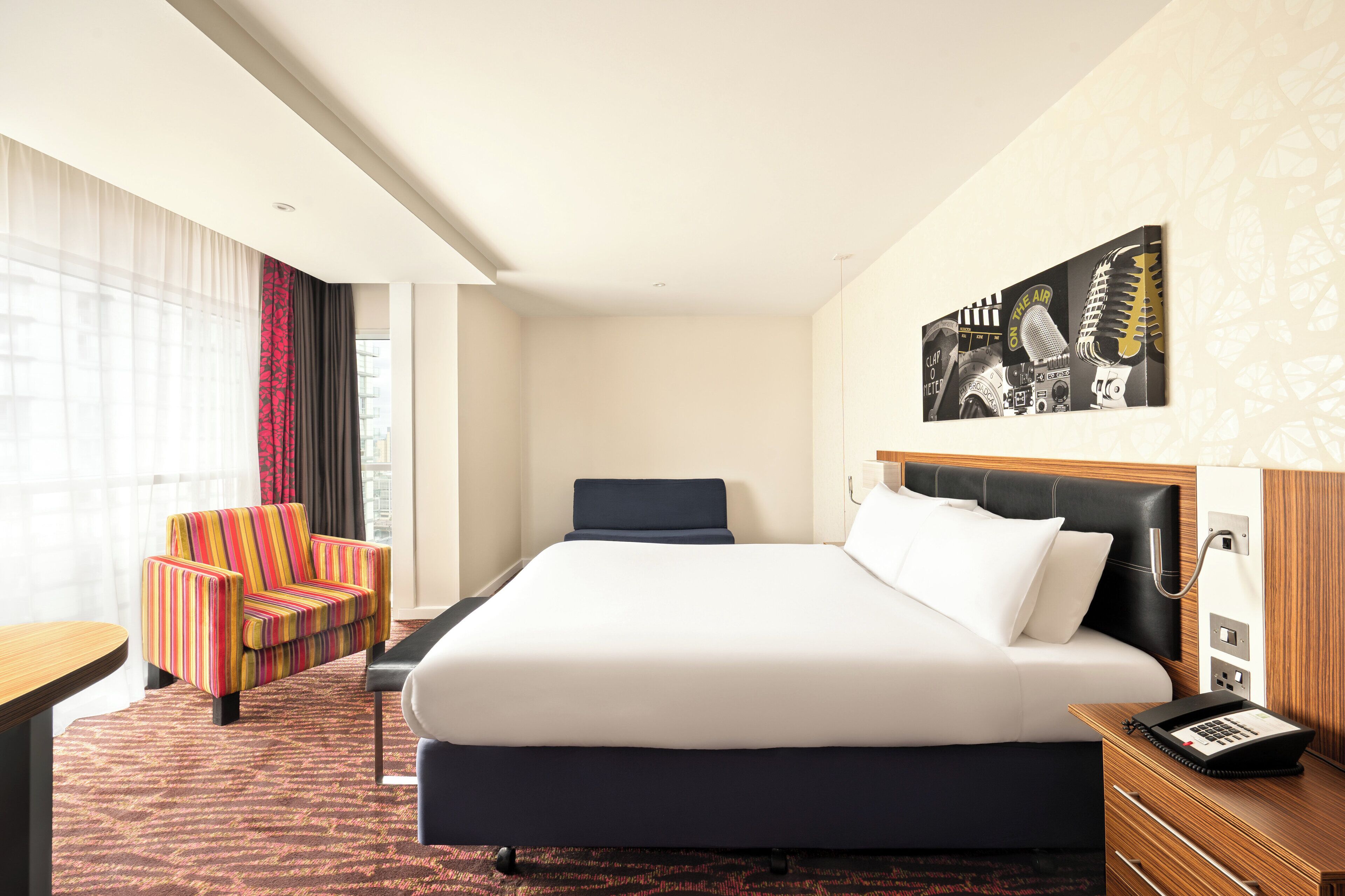 Una habitación de hotel con una cama grande, un escritorio, un sillón colorido y un teléfono en la pared.
