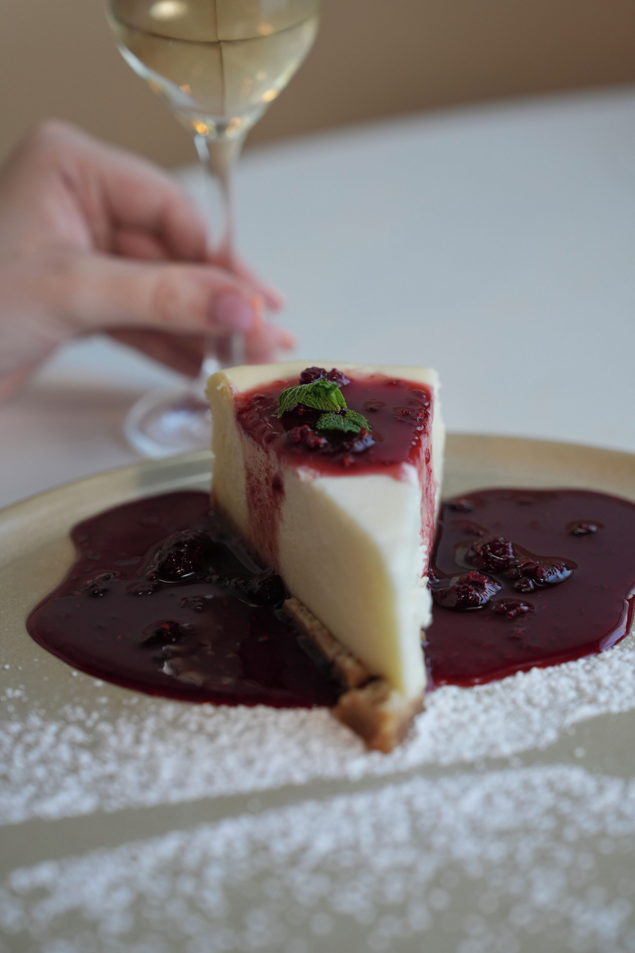 Ein Stück Käsekuchen mit Beerenkompott und einem Minzblatt, bestäubt mit Puderzucker, auf einem Teller mit einer Flüssigkeitslache darum.