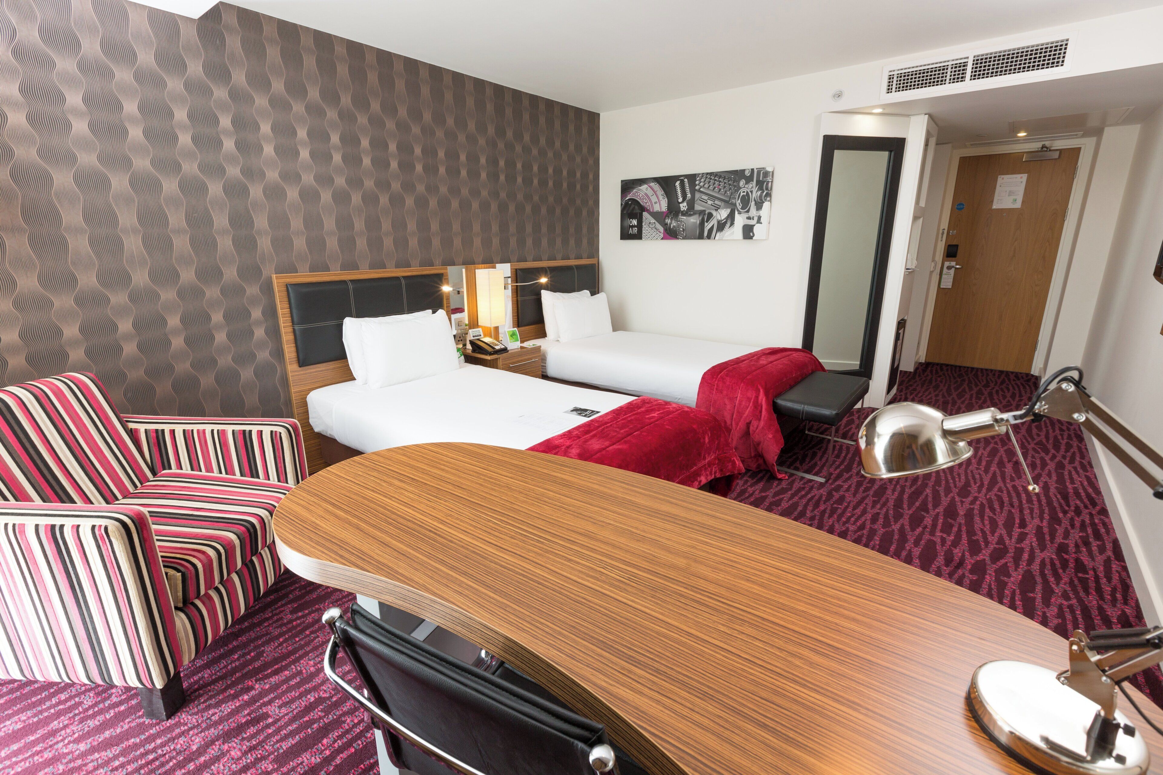 A hotel room with a bed, a desk, a chair, a lamp, and a mirror.