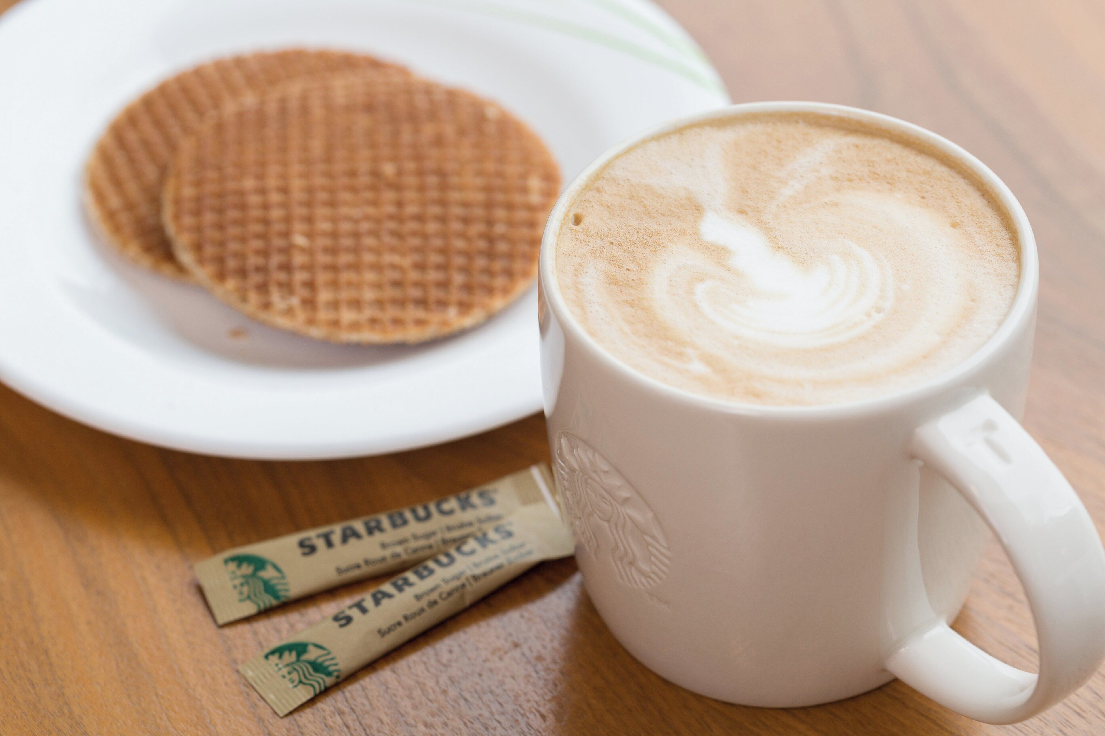 Valkoinen kahvikuppi, jonka pohjalla on latte-kuvio, on puupöydällä. Sen vieressä on lautanen, jossa on vohveli, sekä kaksi Starbucks-kahvipakettia.