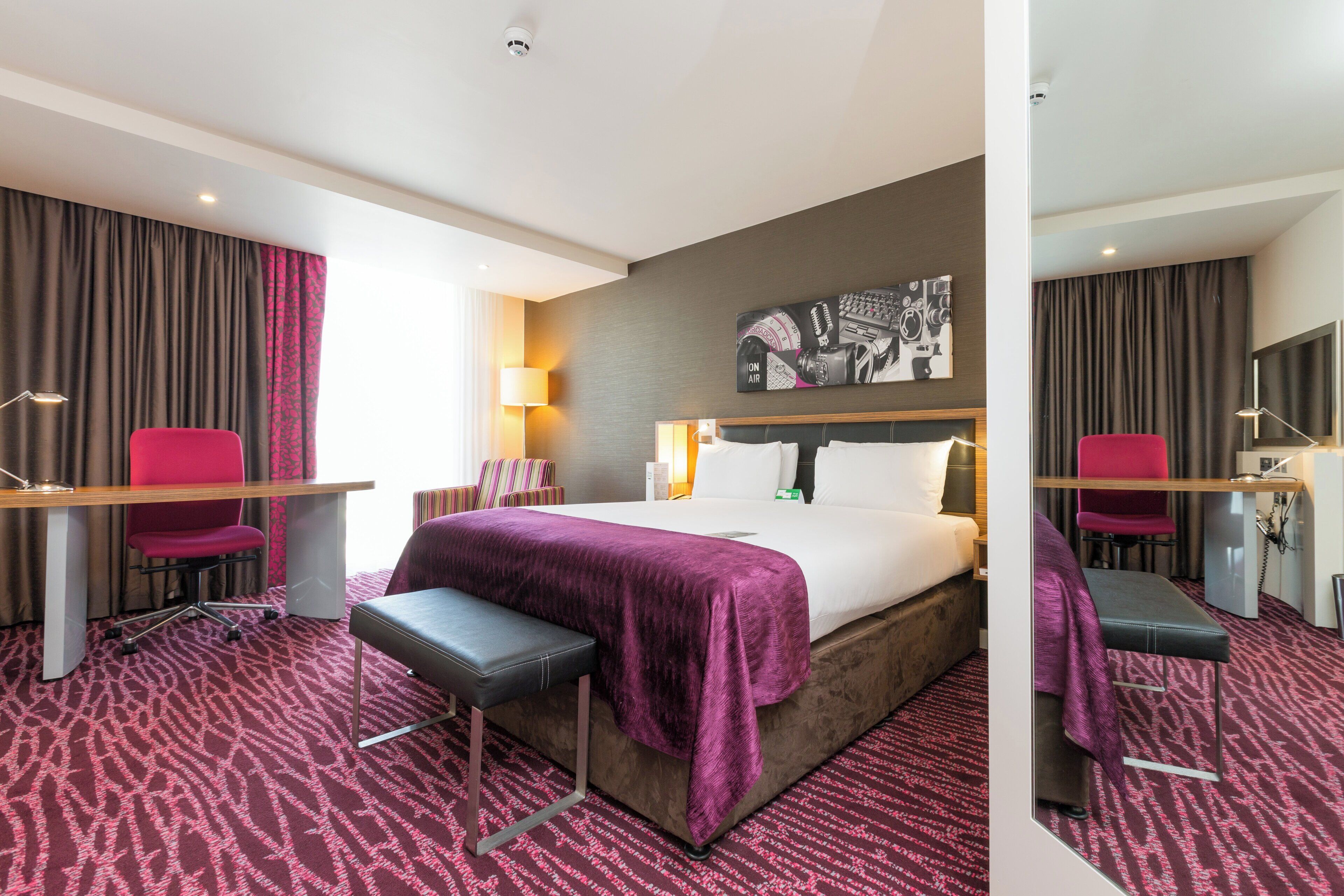 Une chambre d’hôtel équipée d’un lit, d’un bureau, d’une chaise, d’un banc, d’une lampe et d’un téléviseur fixé au mur.
