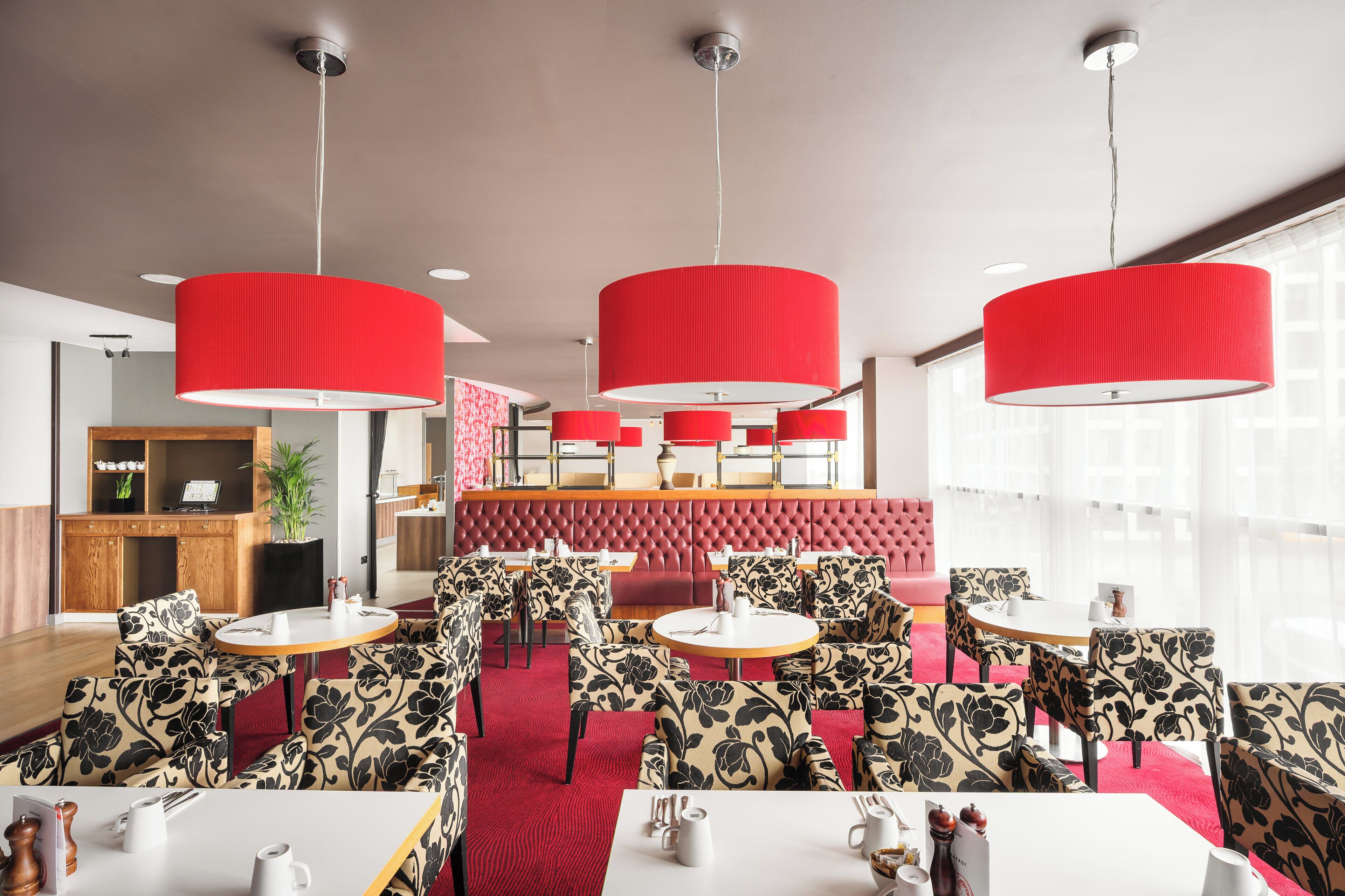 A modern dining area with red upholstered chairs and tables, a red cushioned booth, and pendant lighting.