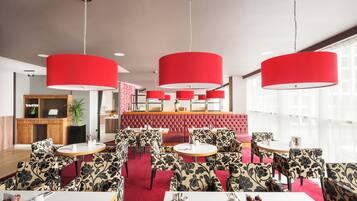 A modern dining area with red upholstered chairs and tables, a red cushioned booth, and pendant lighting.