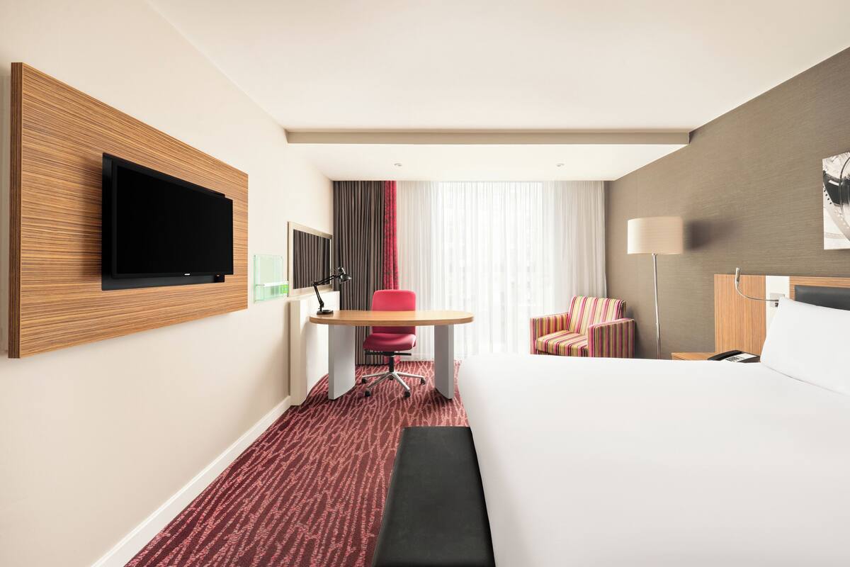 Una habitació d'hotel amb un llit gran, una taula de treball amb una cadira, una televisió penjada a la paret i una finestra amb cortines.