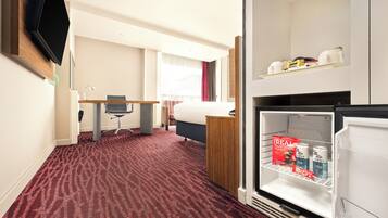 A hotel room with a red carpet, a bed, a desk with a chair, a television mounted on the wall, and a refrigerator stocked with beverages.
