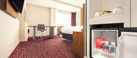 Habitación de hotel con alfombra roja, una cama, un escritorio con silla, televisor montado en la pared y un refrigerador lleno de bebidas.