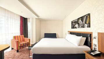 A hotel room with a large bed, a desk, a colorful armchair, and a wall-mounted artwork.