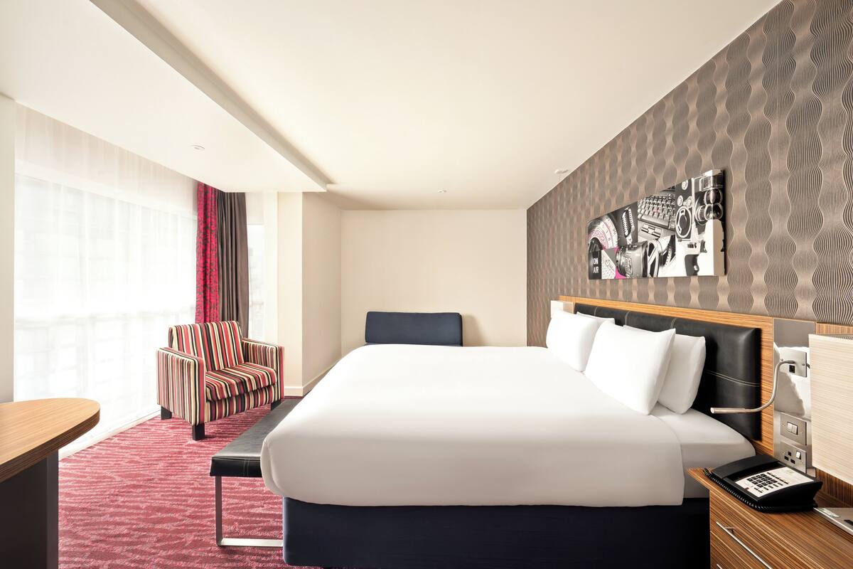 Habitació d'hotel amb un llit gran, una taula de treball, una cadira i una obra d'art penjada a la paret.