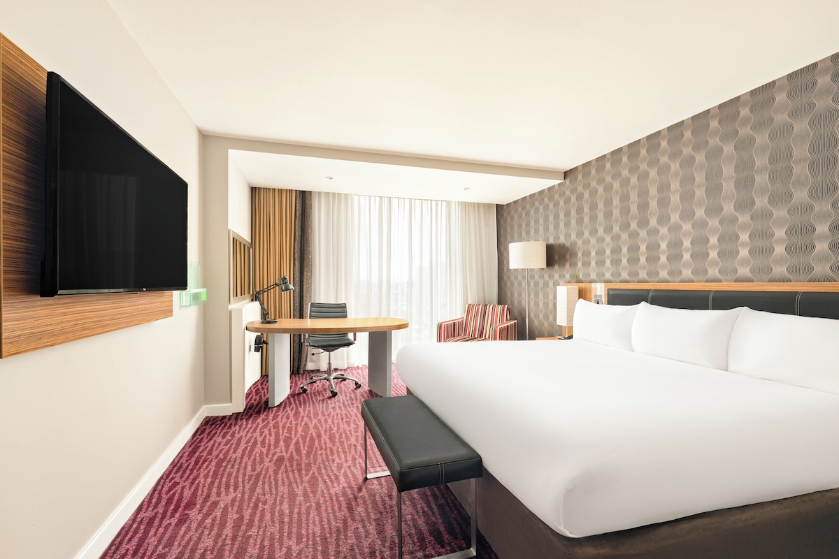 Una habitación de hotel con una cama grande, un escritorio con una silla, un banco, un televisor montado en la pared y un papel tapiz estampado.